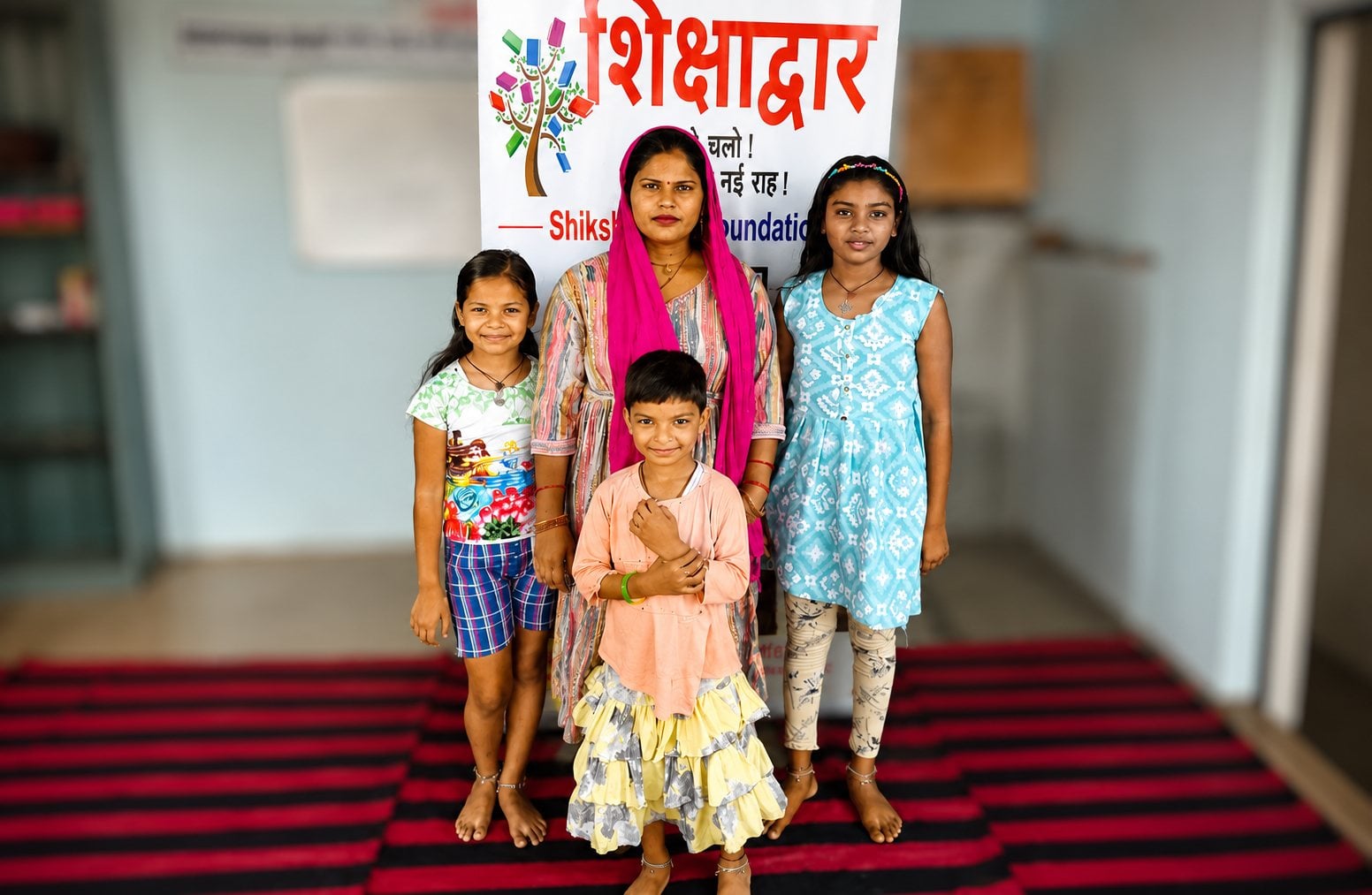 A mother and her children stand together in front of a Shikshadwar Foundation banner