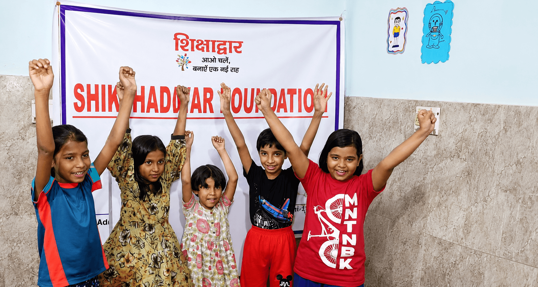 Children cheer with their fists raised in front of the Shikshadwar Foundation banner